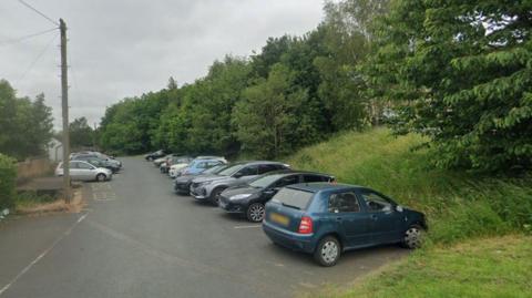 A small car park filled with parked vehicles. Trees and a sloped grass verge sit alongside the car park.