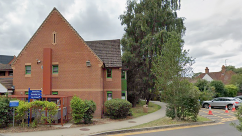 Street view of part of St Ann's Hospital. It is a brick building surrounded by trees and a pathway. To the right of the image a parking space has been coned off. There is a blue sign pointing to Haven Ward which is part of the Psychiatric Intensive Care Unit