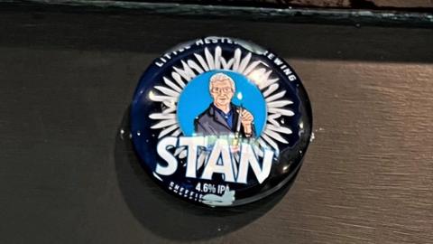 A circular blue badge depicting a cartoon image of a man with grey hair and glasses, Stan Shaw, holding a knife with a circle of assorted blades around him