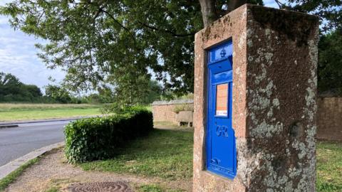 A blue Guernsey post box on a roadside. It is encased in a large stone.