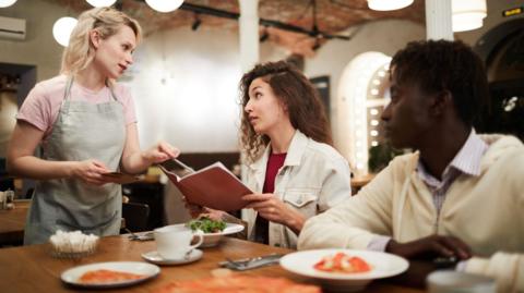 Disgruntled curly-haired young female sitting at restaurant table with boyfriend and reading menu while talking to waitress in modern loft cafe