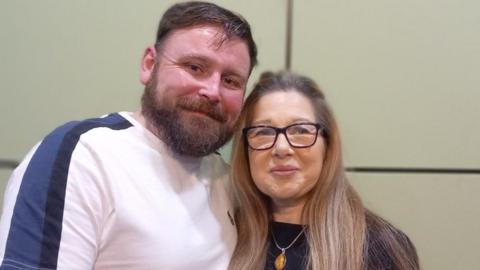 Paul Briggs on the left has dark hair and a beard and is wearing a white and blue t-shift. He is next to partner Viki Smith, who has long hair and is wearing glasses and a necklace.