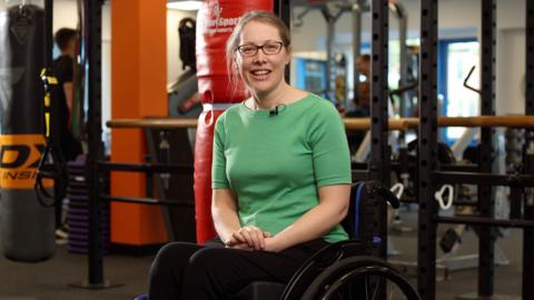 Claire Trivedi, a woman with long blonde hair and glasses wearing a green T-shirt and black trousers, sits in a wheelchair in a gym. Various equipment including punchbags and wall-mounted bars can be seen behind her.