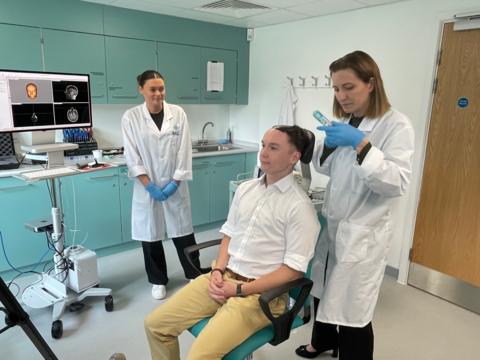 A demonstration with a colleague showing how gel is first placed on the head before an ultrasound transducer is held against the scalp. There are two researchers, with another member of staff demonstrating the process for illustrative purposes.