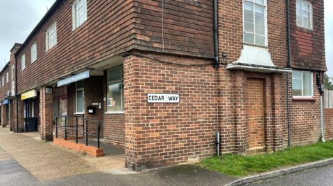 An image of the sign for Cedar Way, Guildford, Surrey. A number of shop units can be seen in the background.