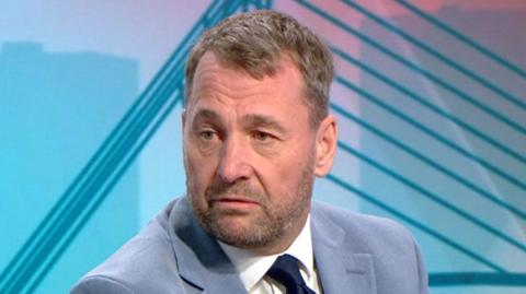Stephen Atkinson sitting in front of a blue striped TV set. He has short greying hair and a short beard and is wearing a grey/blue suit, white shirt and navy tie. He is looking to the left.