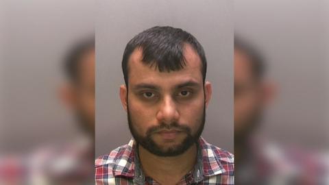 Police custody image of Mohammed Ali he has short dark hair and beard and a checked shirt