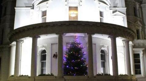 An ornate government office building, which is three-stories with circular pillars around the ground level. The building's white exterior is lit up at night, and a Christmas tree is positioned on the ground level between the pillars. 