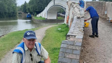 A male volunteer in a blue cap reading 'Canal & River Trust' smiling at the camera. He is crouching down next to the bridge, where other people are working on repairing the bricks. 