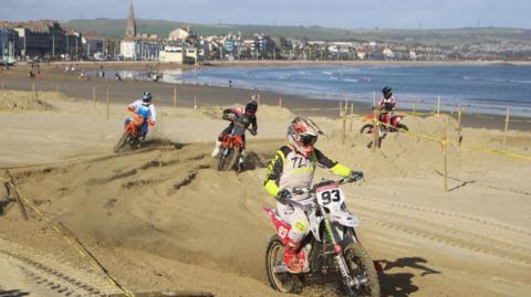 Bikes racing on wet sand with Weymouth town and the sea in the distance. It's a sunny day.