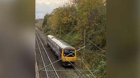 Train stranded on a railway track after winds blew overhead power cables onto the train. The train is surrounded by cables and is next to a fairly steep embankment which is covered in trees and shrubbery.