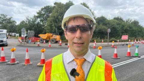 A man in a yellow high vis vest is standing in front of a road that is being worked on. He is wearing a white helmet and dark protective goggles. He is smiling.
There are many cones in the background, as well as a works access sign and an orange digger.