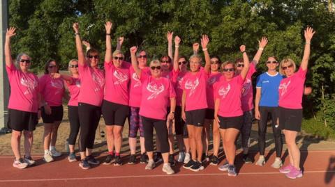 Team Phoenix 2026 stood at running track. All the women are wearing pink t-shirts for their running team and most have their right hand in the air. Everyone is smiling to camera and most are wearing sunglasses.