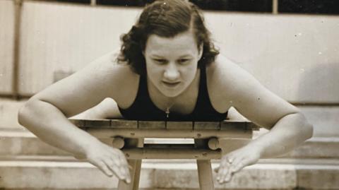 A 1930s black and white picture of a woman lying in a wooden table practising her breaststroke. 