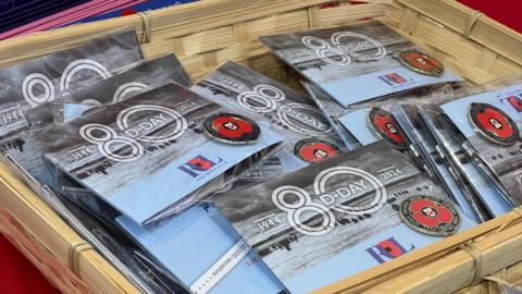 The pins are in a wooden basket on a red table. The poppies have the Jersey flag in the centre and a blue circle around the outside with golden text and the number 80. They are attached to cards which have an image of the D-Day landings on with the dates 1944 and 2024 on either side of the number 80 which had D-Day writted on the inside of the 0.