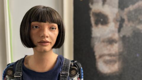 Ai-Da is a humanoid robot artist. Shown with her picture of Alan Turing in the background.