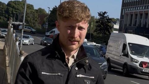Matthew Stewart leaves court in Carlisle, outside Carlisle Magistrate's Court. He has blonde/strawberry blonde coloured short curly hair and stubble. He is wearing a black biker jacket which is large silver zips. 
