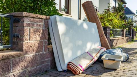 A mattress, rolled-up rugs and a washing up bucket left on a pavement outside a home.