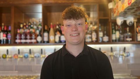Joshua is wearing a black shirt and is behind the bar. 