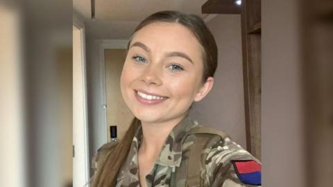 A young woman wearing army camouflage is smiling in a selfie style photo.  She has her hair pulled back into a ponytail, which is sat over her shoulder. There is a red and blue patch on the top of her sleeve.