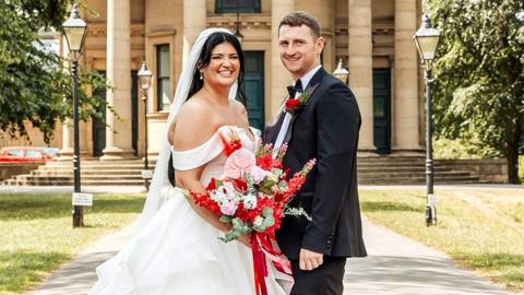 A woman in a white wedding dress stands with a man in a suit. She holds a bouquet of red and pink flowers with a red ribbon. They stand at the pathway to a round church, metal ornate gates are open next to them and the path lined with traditional street lights. To the right and left is a lawn with large trees.