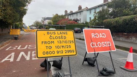 A yellow and black sign saying the bus lane is closed until 1o/11/25 next to a red and white sign saying road closed. The lae to the left is marked as a bus lane and there is a traffic cone to the right.