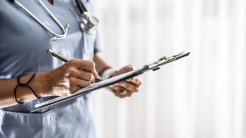 Close up of a medical professional writing on a clip board. We cannot see her face.