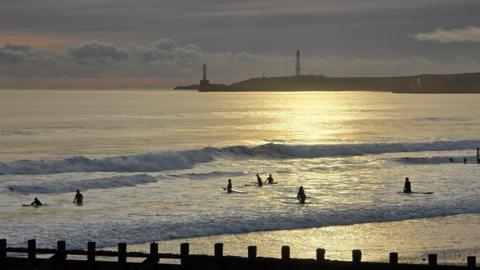 Surfers go out into the sea which is golden and blue reflecting the rising sun
