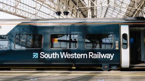 Dark blue train with white detailing stopped on a platform at a station a door on its far right is open.