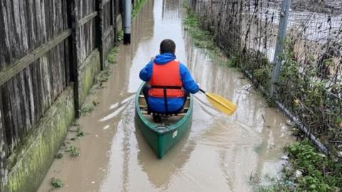 A man with his back to the camera, travelling through floodwater. He is wearing a life jacket, and blue coat. His canoe is green and his oars are yellow. 