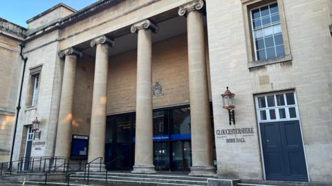 The frontage of Gloucestershire County Council's Shire Hall. The front of the building has four floor to ceiling concrete pillars at the top of the steps, with the entrance doors behind them. The name of the building is written on both sides of the pillars.
The sky is visible at the top of the photo, which is bright blue.