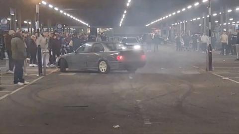 A grey car appearing to spin around doing a trick in a car park while crowds of people watch