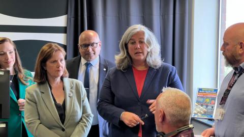 A woman in a navy blazer and long grey hair speaking to a man sat down in front of a computer 