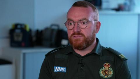 James Vernon, who has short ginger hair, a ginger beard and round framed glasses, sits on a chair and speaks to the camera wearing his green North West Ambulance uniform.