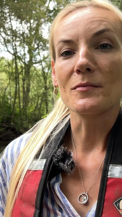 A woman wearing a life jacket is standing in a stream, she has blonde hair.