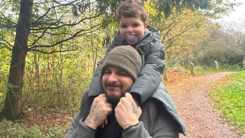 A man with a short beard wearing a grey beanie smiles towards the camera. His young son is sitting on his shoulders, he is wearing a grey coat and has a purple dummy in his mouth. They appear to be in a park or on a woodland trail, there are trees and a dirt path behind them