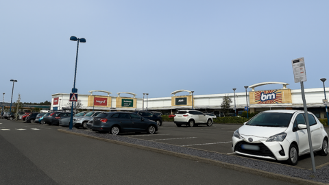 The image shows a retail and leisure park car park on a bright, clear day. The sky is cloudless and light blue, suggesting good weather and daytime conditions. The scene appears calm and orderly, with no visible crowds.