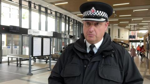 Supt Dave Henthorne is standing inside the bus interchange. People can be seen milling around in the background. He is wearing a black police hat and jacket. 