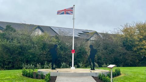 A concrete plinth with two metal black silhouettes of soldiers, a flagpole between them with a union jack flag and a poppy tied to it halfway down. Small shurbs have been planted along the path and around the plinth. All surrounded by grass.