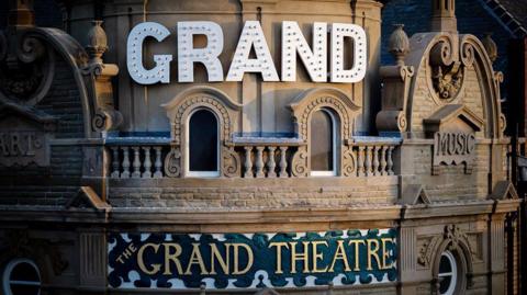 A close-up of the white illuminated sign on the theatre that says "GRAND". Underneath is a smaller gold and green sign that says "The Grand Theatre" on the front of a round stone frontage