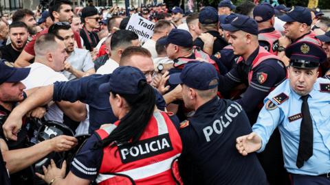 A crowd of demonstrators clash with police during a protest. Officers in red and blue uniforms which say 'POLICIA' hold back male protesters.
