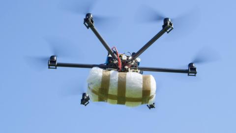 A multi-rotor drone flying in the sky while carrying a package suspended beneath its frame. The photograph is taken from below the drone, looking upward toward a clear blue sky.