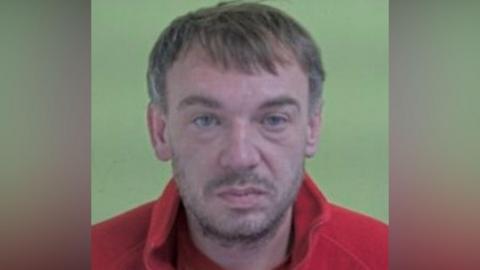 Police photo of Ross Beggs-Cairney, who has short brown hair and stubble and is wearing a red fleece top.
