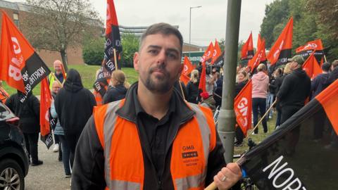 Dozens of picketers are waving flags in the background whilst a man, with short brown hair and a beard, looks into the camera. The flags are coloured orange and black, with GMB branding on them.