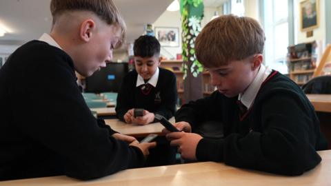 Three boys in a library in school uniform. They sit facing each other using old Nokia "brick phones".