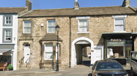 The register office in Barnard Castle is within a two-storey beige stone building with white windows. It is next to an arched doorway, a cafe and a shop.