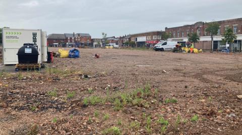 A view of the empty Royal Arcade site. It is a large swathe of dry, exposed soil with a few tufts of grass. There are shops and buildings around the permiter. There is a white van and some equipment on site, including a metal containers and some large materials bags.
