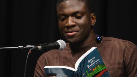 A man reads into a microphone whole holding a book. He is wearing a brown striped shirt