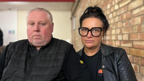 Mark and Caroline Willgoose - a middle-aged couple staring into the camera. Mr Willgoose has short grey hair, while Mrs Willgoose has black hair and thick black-rimmed spectacles. Both are wearing black coats and are sat by a brick wall.