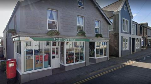 Aspatria's former Post Office on King Street. It is a grey building with windows running along the front and a Post Office sign above the entrance. A red post box is to the right of the building.